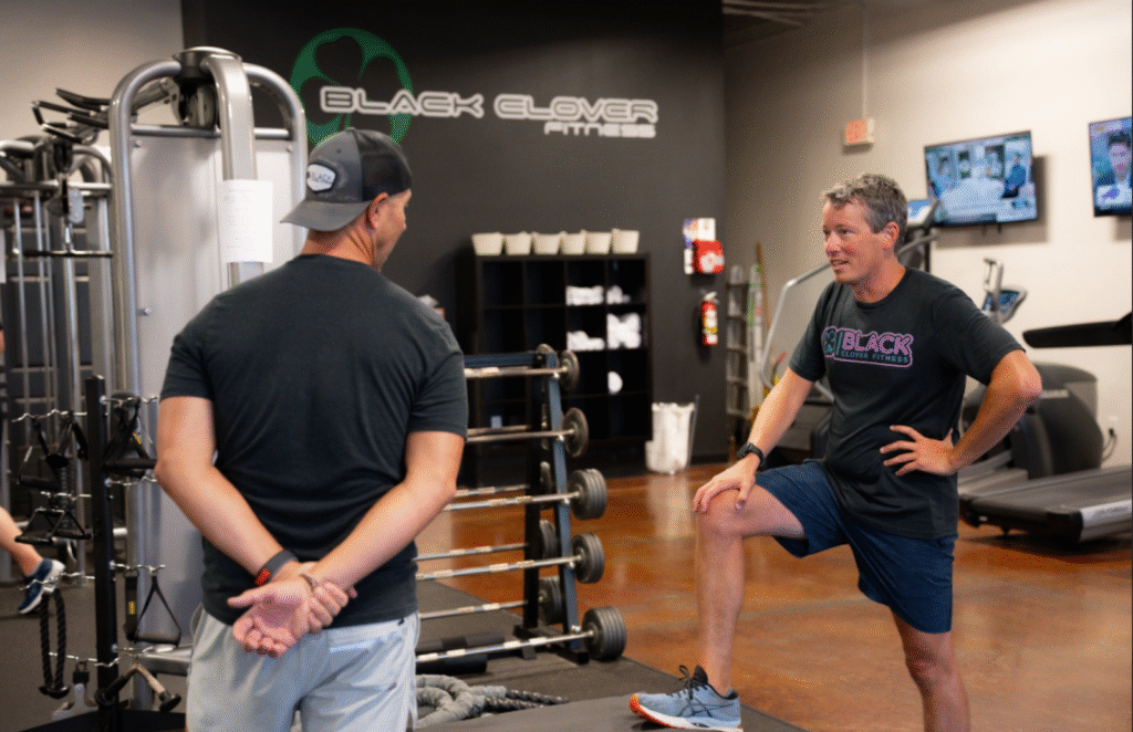 Two men engaged in a fitness discussion at Black Clover Fitness, one demonstrating a stretching exercise, emphasizing community support and personalized training for GLP-1 users in Omaha.
