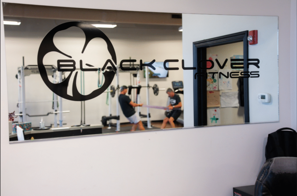 Black Clover Fitness logo on a mirror with two individuals engaged in strength training exercises in a gym setting, emphasizing resistance training for GLP-1 users.