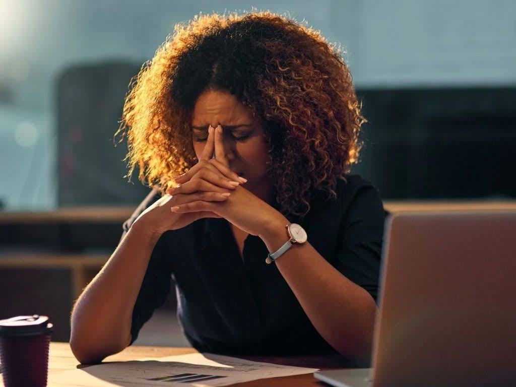 Woman with curly hair showing signs of stress, sitting at a desk with a laptop and papers, reflecting the challenges of maintaining health and wellness amidst weight loss efforts.